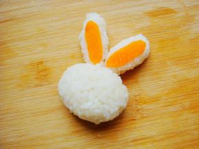 先来做小兔子：蒸好的米饭放入保鲜膜里团成一个圆球做兔子头部。团两个水滴状的饭团做耳朵，贴上长条状的胡萝卜片点缀下。