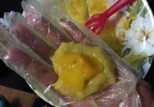 准备一碗水，一个一次性手套，这个方法是小编试过最好用的了，戴好手套，抹一点点水，然后抓一点点糯米面注意烫伤，然后中间塞一个芒果肉，然后用勺背压一压这个芒果肉，压进糯米面里，然后右手也要少沾点水，因为糯米面实在太黏了，像包包子一样包好，左手倒右手，右手倒左手，撮圆了