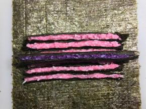 尽量把紫色搓圆。底下铺一张新的海苔，铺好粉色的条，再把紫色条放中间，卷起来。