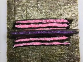 尽量把紫色搓圆。底下铺一张新的海苔，铺好粉色的条，再把紫色条放中间，卷起来。
