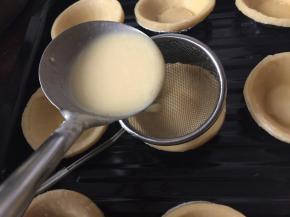 将蛋挞皮放入烤盘里，通过网筛将蛋挞液勺入蛋挞皮里