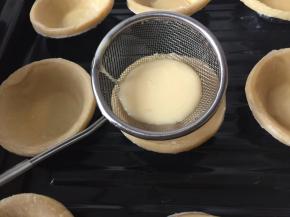 将蛋挞皮放入烤盘里，通过网筛将蛋挞液勺入蛋挞皮里