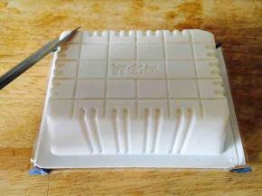 如何能完整地取出内脂豆呢？我有小妙招，首先轻轻拍打内脂豆腐的正反两面，各拍打5下，然后用双手轻轻在盒子四周，往里捏两下，最后再用剪子在盒子的反面剪两个小角，这两个小角是斜对称的。