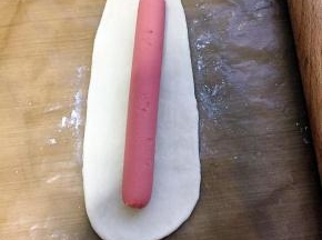 面团排气后用擀面杖擀成披萨盘大的面皮，用刮刀刮掉多余的面皮，多出来的面皮用来做花边，擀成宽宽的长条，放上火腿，包起来