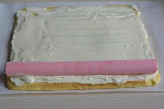 将奶油奶酪糊均匀地抹在蛋糕片上，开始的部位可以厚一点，结尾的部位抹薄薄的一层；将冻好的粉色奶酪夹心从模具中取出，速度要快噢；取合适的长度铺在蛋糕片的一端；从下向上卷起，然后用油纸包裹好，再略微用力按一下，使夹心能紧密贴合