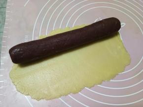 待红色面团冻硬了，把原色面团下面铺好保鲜膜，然后在保鲜膜上面擀成长方形大薄片，尺寸可以包裹住红色圆柱面团。借助保鲜膜，像卷蛋糕卷一样的手法把原色面团包裹住红色面团，并且隔着保鲜膜轻轻攥一下，让两种颜色的面团可以粘贴紧实。