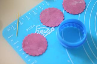 取一块白的翻糖膏加入淡紫色素，并擀成薄片，用纸杯大小的饼干模切割出一个带花纹的圆形。