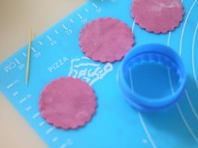 取一块白的翻糖膏加入淡紫色素，并擀成薄片，用纸杯大小的饼干模切割出一个带花纹的圆形。