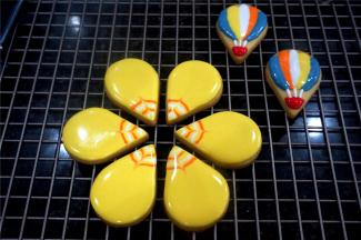 取6个水滴状的饼干，先填上黄色打底，然后从里至外，分别用白色，橙色，白色，橙色画弧线，画好后，用牙签从水滴饼干尖的中部往外拉线，形成花纹。另外取两个水滴饼干，用不同颜色的糖霜画出热气球的图案，画好后放置一旁晾干