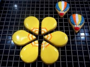 取6个水滴状的饼干，先填上黄色打底，然后从里至外，分别用白色，橙色，白色，橙色画弧线，画好后，用牙签从水滴饼干尖的中部往外拉线，形成花纹。另外取两个水滴饼干，用不同颜色的糖霜画出热气球的图案，画好后放置一旁晾干