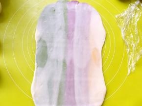 用擀面杖顺着彩色油酥皮纹路擀成长条，之后卷起来，盖保鲜膜松弛15分钟，擀的过程如果有气泡，可以用牙签扎破，再捏紧