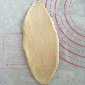 取一个圆面团，用排气擀面棍将面团擀成23CM长的椭圆形。右手用刮板将面团铲起，左手将面团翻面