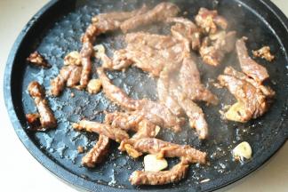 电饼铛开下盘，下入色拉油烧热并爆香蒜片辣椒。下入腌好的牛肉条翻炒至变色。