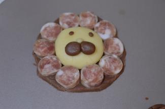 再取两个可可面团揉圆，形成狮子的鼻子，同时也把眼睛贴上，并把香肠旁边多余的可可面团去掉。