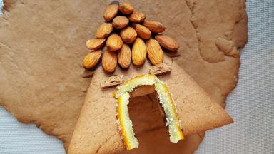 12．饼干放凉后，大杏仁蘸融化后的黑巧克力粘在房屋上做瓦片，糖渍橙皮做门框，多余的饼干切两片做窗户，室温放置使巧克力凝固。