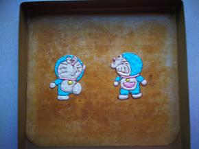 将白纸铺在不粘烤盘上，上面铺放一张油布，用咖啡色在油布上描出机器猫的形状，用其他的颜色进行填充。全部做好后放入冰箱冷藏保存。