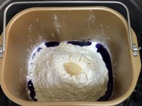 在面包机桶内加入蝶豆花水，细砂糖和盐对角放入，接着倒入全麦面粉和高筋面粉，最后在面粉上挖个洞，加入酵母粉（除黄油以外的材料，按照先液体后粉类的顺序），启动面包机和面程序。