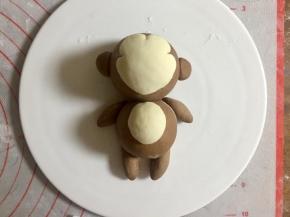 把12步骤的可可小面团揉成橄榄的形状，一端沾点水贴在小猴子四肢的部位；. 再搓两个可可面团小圆做猴子的耳朵，沾水粘在头部两侧；然后再取一小团白色面团，揉圆按扁，沾水贴在身体的中间，做猴子的肚皮。