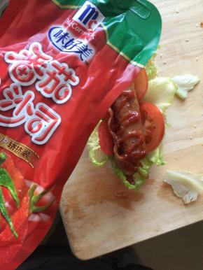 然后将做好的面包在中间切开，塞入生菜、番茄片、培根和热狗肠、挤入番茄酱