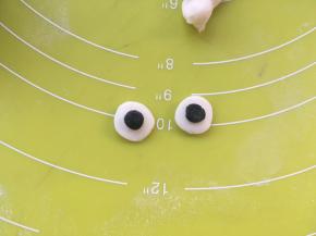 取两小份白色面团搓圆压扁，再取两小份黑色面团搓圆压扁，点一点点水黏在白色面团中间，就形成了蓝精灵的眼睛