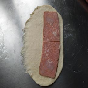 擀成椭圆形一边用手压平底边如图，放一片培根，放整条的，我是因为家里没有整条的培根。边上留1-2cm的空隙，撒少许黑胡椒，从一边卷起，收口向下排入烤盘。