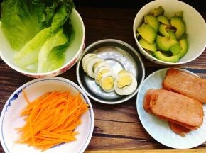 先将午餐肉切片，然后平底锅里倒入少许油，将午餐肉煎至两面稍黄，装入盘子里备用，接着将生菜洗净沥干水，胡萝卜洗净去皮后切丝，鸡蛋煮熟后切片，牛油果去掉外皮、去核以后切片，胡萝卜丝可以用海盐和黑胡椒碎调味。
