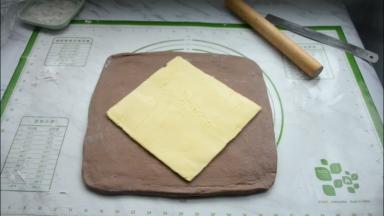 将松弛好的面团取出，在硅胶垫上撒些面粉放粘连，再把面团擀成比黄油片稍大的面片，把黄油片放在中央。