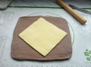 将松弛好的面团取出，在硅胶垫上撒些面粉放粘连，再把面团擀成比黄油片稍大的面片，把黄油片放在中央。