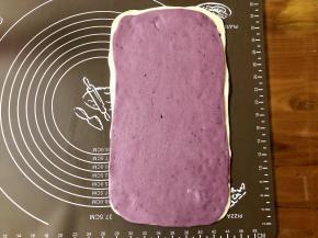 然后在白色面片上喷一些水，把紫色面片叠上去，用擀面杖轻轻擀一擀，让两张面皮粘合在一起。
