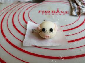 将耳朵粘在相应的位置，要想粘得更牢固，可以沾点水。黑色面团揪出一点点，做出眼睛。再揪出一点点粉色面团，做鼻子，用牙签戳出两个小孔。粉色面团搓出细细的长条，切成5份小段，粘在小猪后面，做猪尾巴