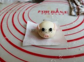 将耳朵粘在相应的位置，要想粘得更牢固，可以沾点水。黑色面团揪出一点点，做出眼睛。再揪出一点点粉色面团，做鼻子，用牙签戳出两个小孔。粉色面团搓出细细的长条，切成5份小段，粘在小猪后面，做猪尾巴