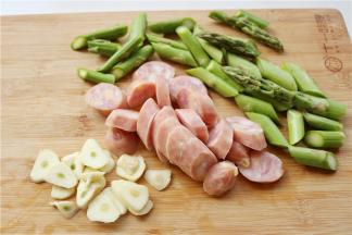 把配菜处理一下，芦笋去除老根，切成段，芝士香肠切成片，大蒜也切成片。