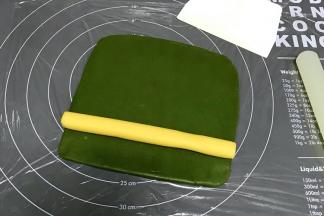 在硅胶垫上铺一张保鲜膜，把绿色面团擀成18*18cm的方形面片，放上原色长柱面团。