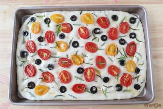 再刷上一层橄榄油，放上油浸小番茄作为装饰，将黑橄榄切片放上去，最后撒上迷迭香和黑胡椒粉。